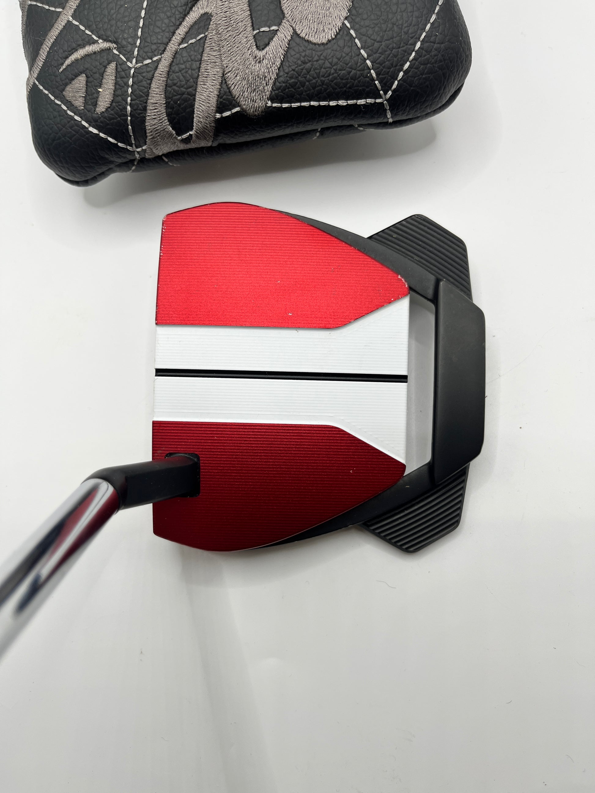 Close-up of a golf putter with red and black head and white blade on a light gray background.